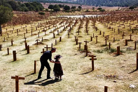 Un hombre y un niño observan un vasto cementerio sad hill, con hileras de cruces de madera bajo un cielo despejado.