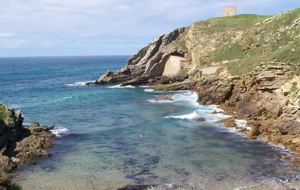 Costa rocosa y mar turquesa en Santillana del Mar, Santander. Una pequeña ermita se esconde en la roca y una torre vigía corona la colina.